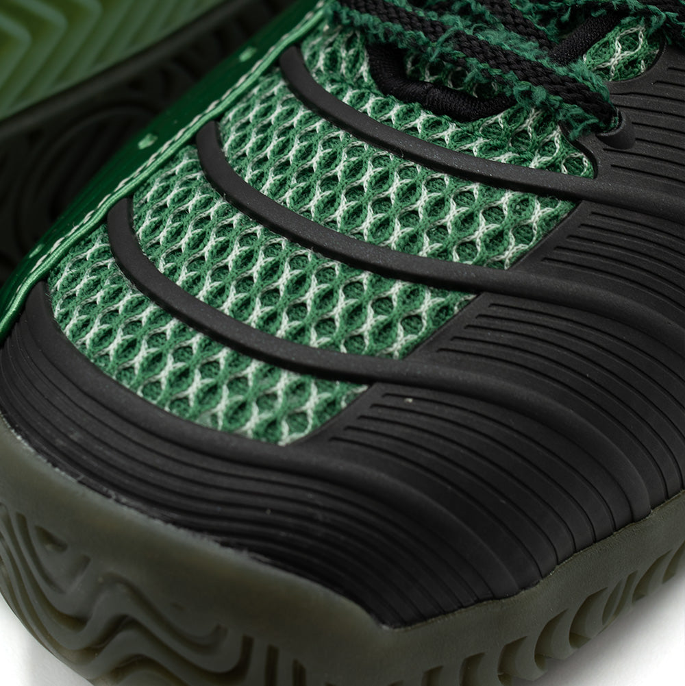 Detailed macro perspective of the forefoot on the adidas x Brain Dead Barricade V, focusing on the dark green open-structure mesh overlaid with grooved black rubber flex guards and the rugged wrapped toe bumper.
