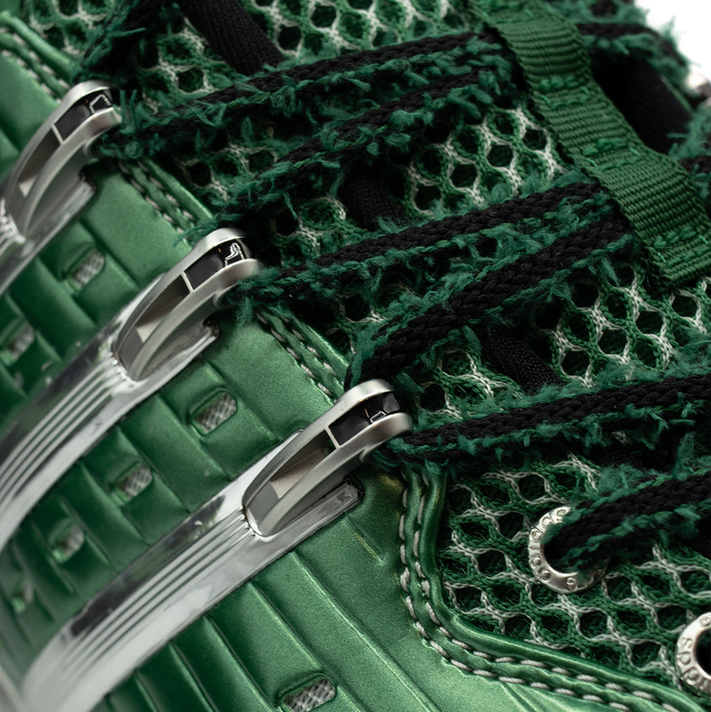 Close-up studio perspective of the lacing system on the adidas x Brain Dead Barricade V, showing the polished chrome Three Stripes cage extending into eyelet anchors, metallic eyelets stamped with "adidas", and two-tone textured woven laces over green mesh.