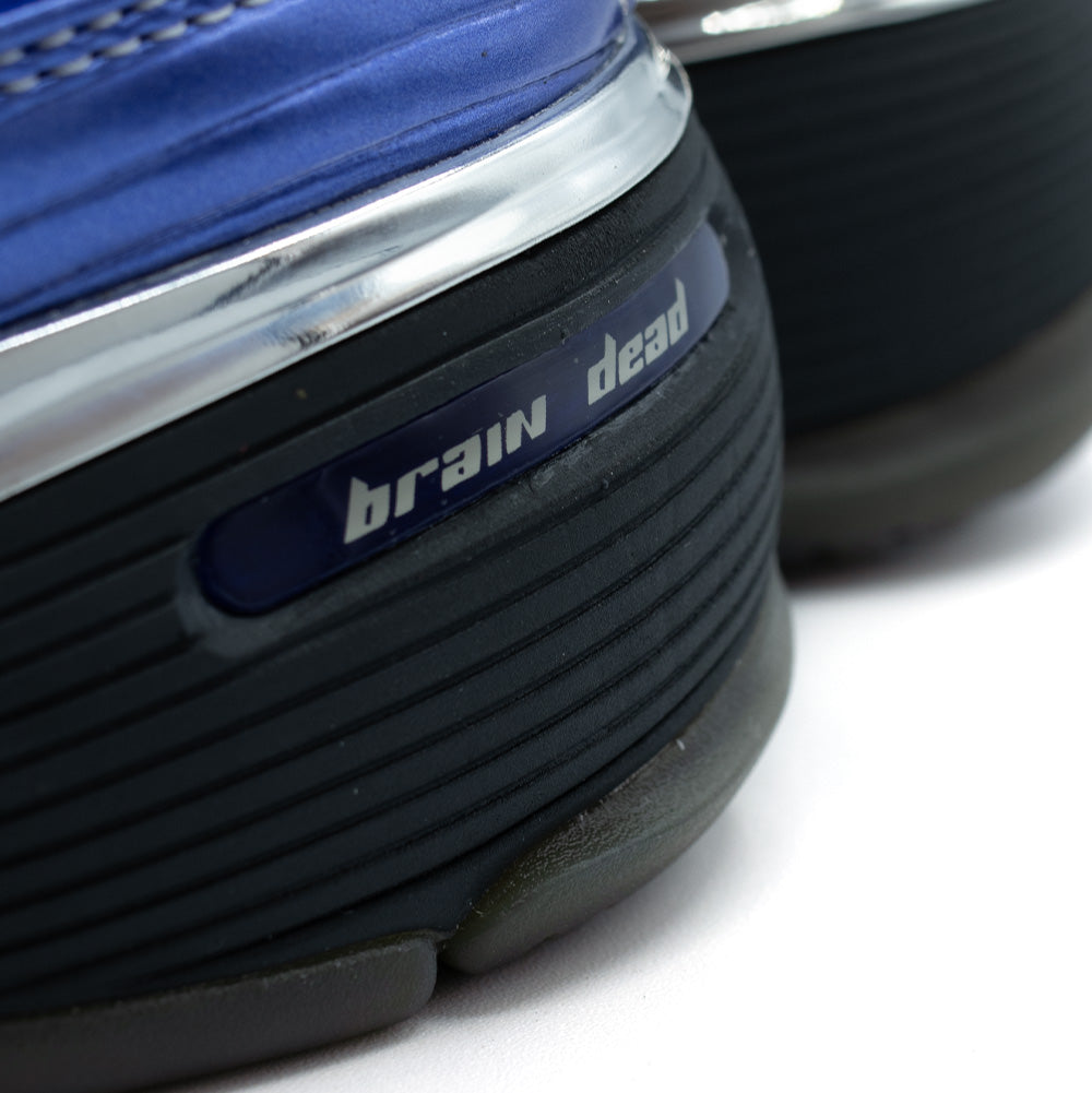 Extreme macro close-up of the lateral rear heel on the adidas x Brain Dead Barricade V sneaker in Mystery Ink, highlighting the horizontally ribbed carbon black midsole chassis inset with a glossy navy blue oval badge featuring silver "brain dead" lowercase branding, framed by a high-shine chrome metallic trim and the ribbed royal blue synthetic upper with tonal stitching.