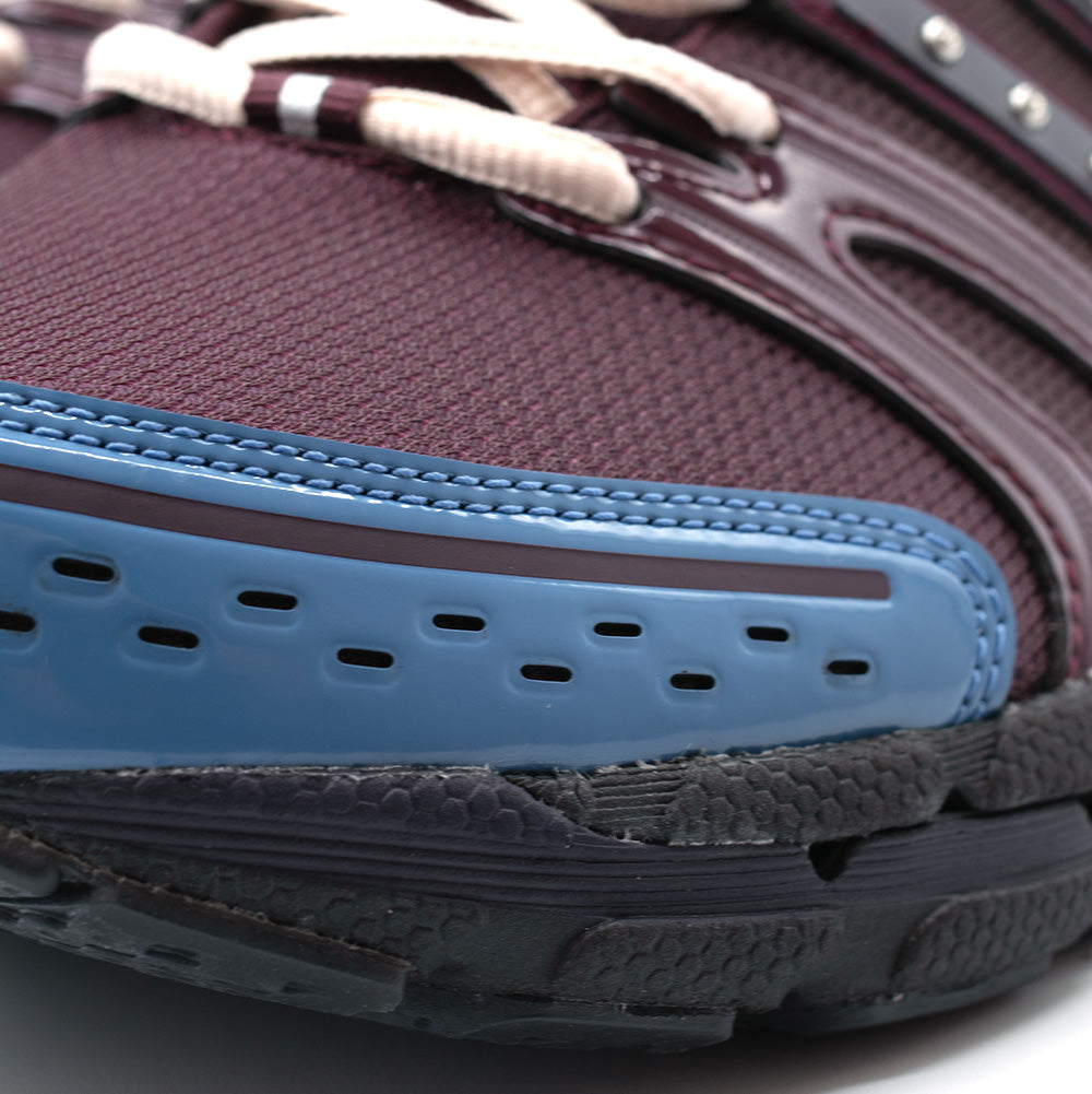 Close-up studio perspective of the forefoot on the Adidas X Hardies Adistar Control 5, showing the periwinkle-blue perforated synthetic toe cap, glossy maroon mudguard trim, breathable dark mesh, and textured black foam midsole wall.