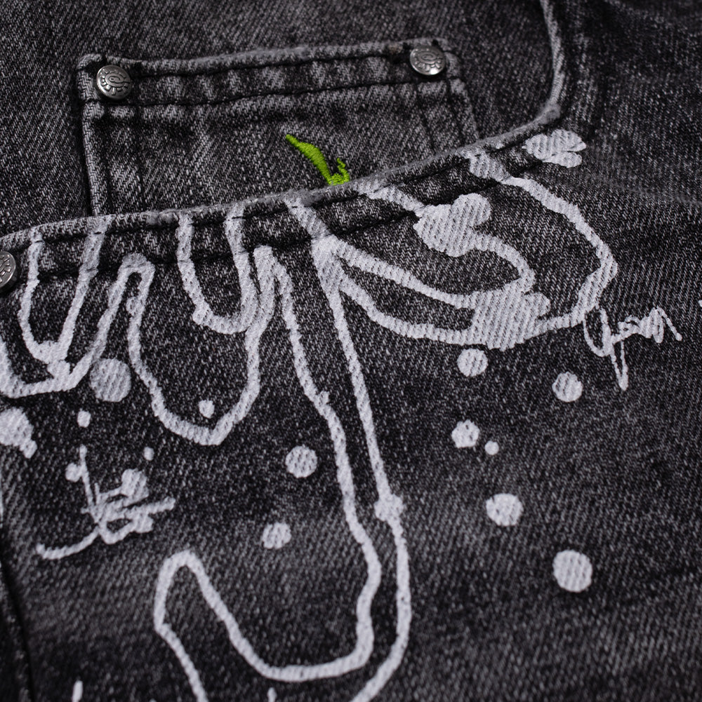 Detailed macro close-up of the front right pocket on the washed black Always Waza Jeans, highlighting the dripping white street-art screen print, custom silver metallic rivets embossed with the @sun logo, and a vibrant lime green embroidered silhouette of New Zealand positioned inside the coin pocket.