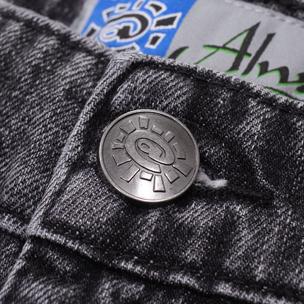 Macro studio close-up of the waistband closure on the Always Waza Jeans in washed black, focusing on the gunmetal-finish shank button debossed with the signature Always @sun emblem, paired with black denim twill and tonal reinforcement stitching.