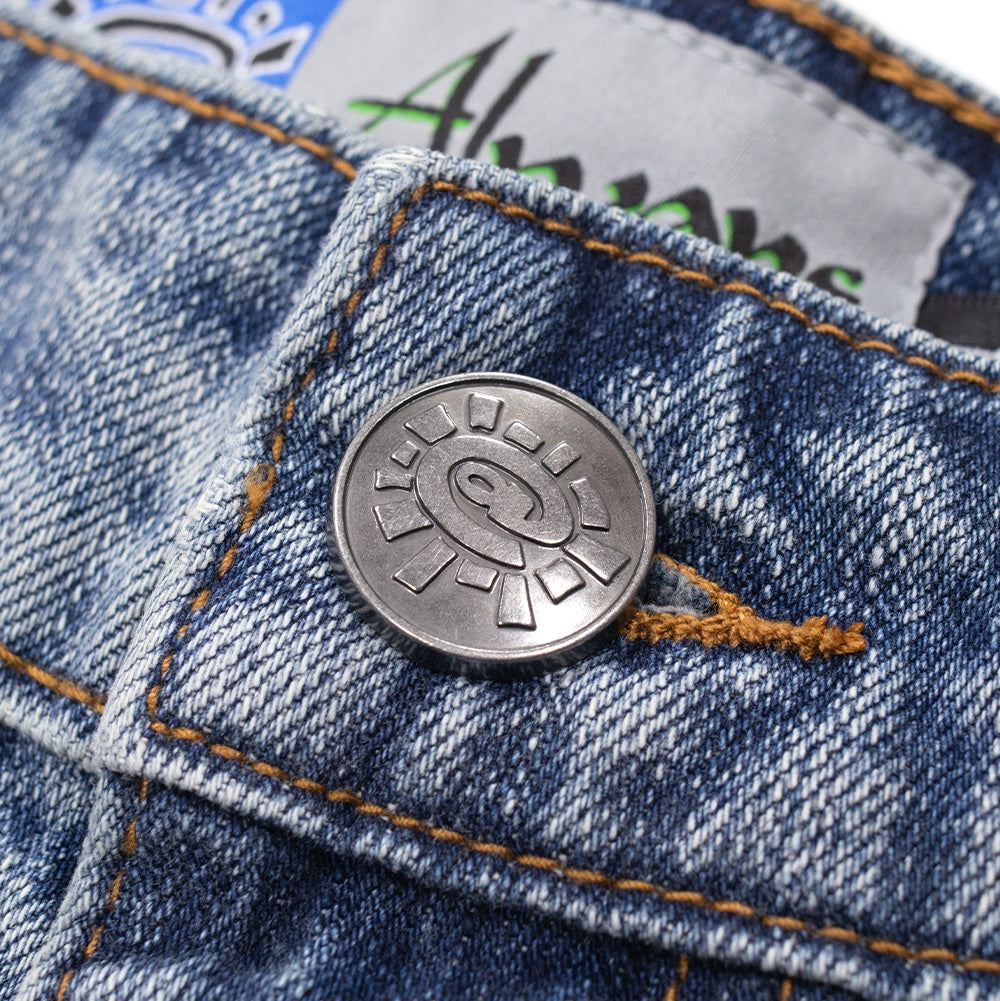 Macro studio close-up of the front waistband closure on the Always Waza Jeans in washed light blue, focusing on the dark antique silver metal shank button debossed with the signature Always @sun emblem, set against contrasting tan thread buttonhole stitching and textured twill denim.
