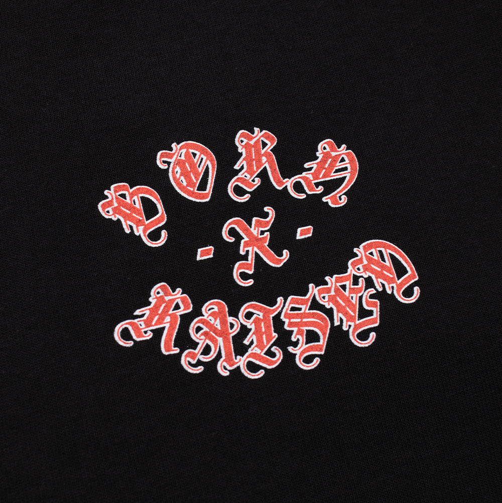 Detailed macro studio close-up of the graphic print on the Born X Raised Gothic Rocker T-Shirt, highlighting the textured scarlet red ink filling the intricate flourishes of the Old English Gothic letters outlined in clean white, spelling out "BORN - X - RAISED" against the deep black cotton fabric.