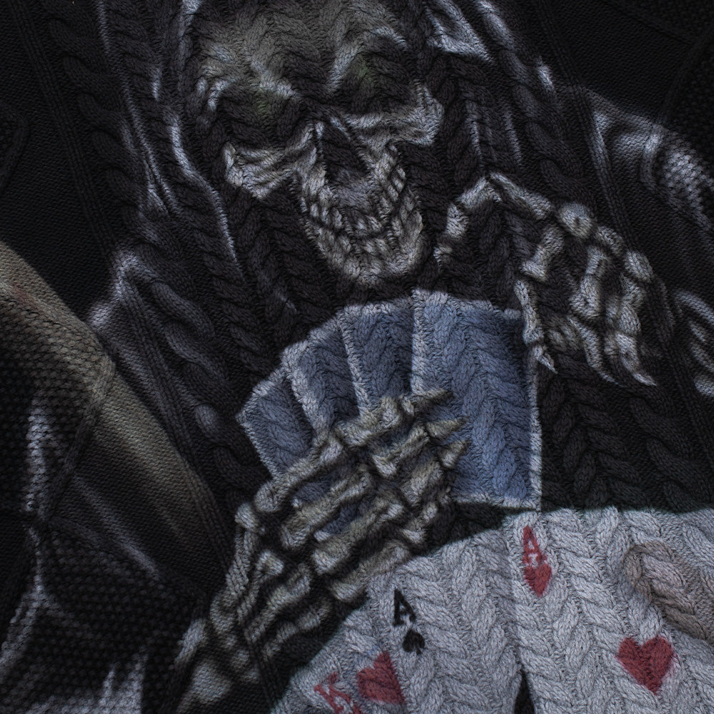 Detailed macro perspective of the front graphic on the Born X Raised Two Pair Cable Knit Sweater, capturing the high-contrast printed artwork of the hooded skull Grim Reaper with glowing sockets, holding a hand of poker cards including the Ace of Spades and King of Hearts across the raised cable yarn texture.