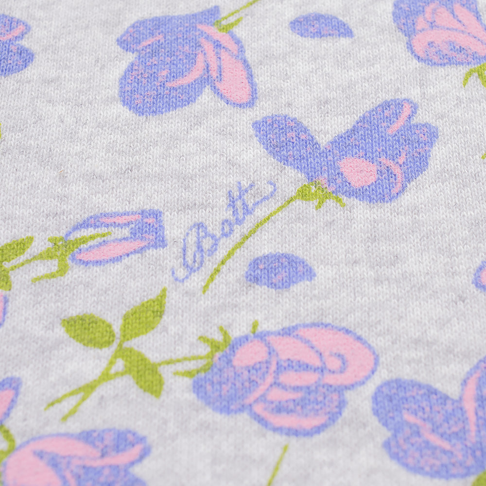 Extreme macro close-up of the all-over botanical print on the BoTT Flower Zip Hoodie, focusing on the delicate pink and periwinkle blue rose petal artwork, textured grey melange cotton-poly fleece knit, and the subtle blue cursive "BoTT" signature branding integrated seamlessly along a flower stem.