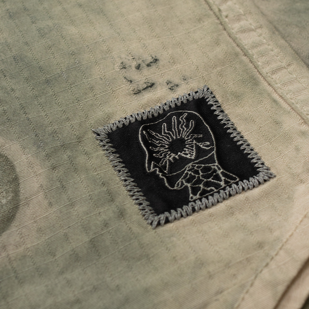Close-up shot of the lower left front hemline on the Brain Dead Algae Shirt Jacket, focusing on a square black fabric brand patch featuring the white embroidered Brain Dead profile head logo with radiating burst graphics, secured by thick decorative grey zigzag edge stitching.
