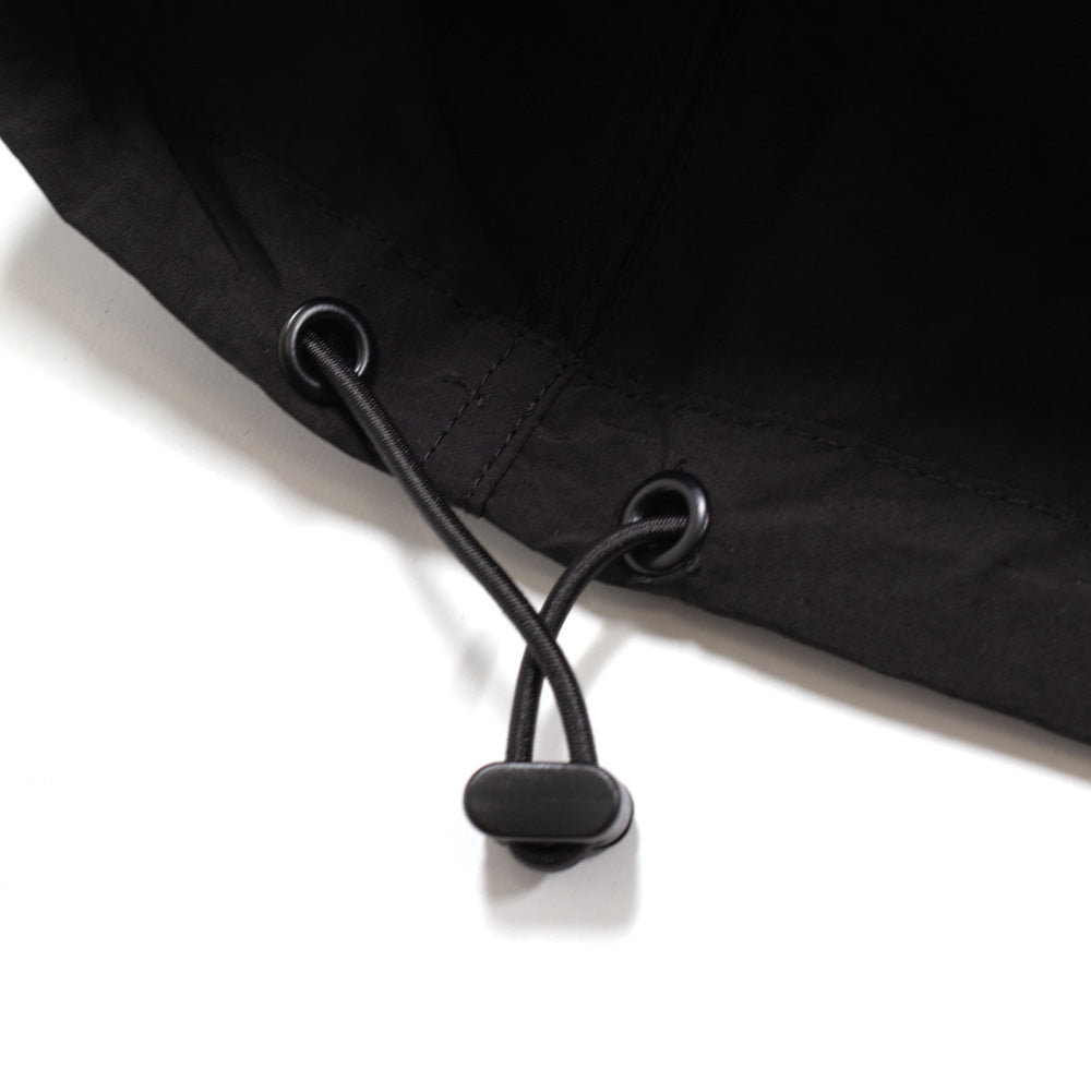 Macro studio shot of the bottom hem cinching mechanism on the Brain Dead Dolman Sleeve Jacket, highlighting two black matte metal eyelets, a round elastic bungee drawcord, and a contoured black plastic barrel cord lock stopper.
