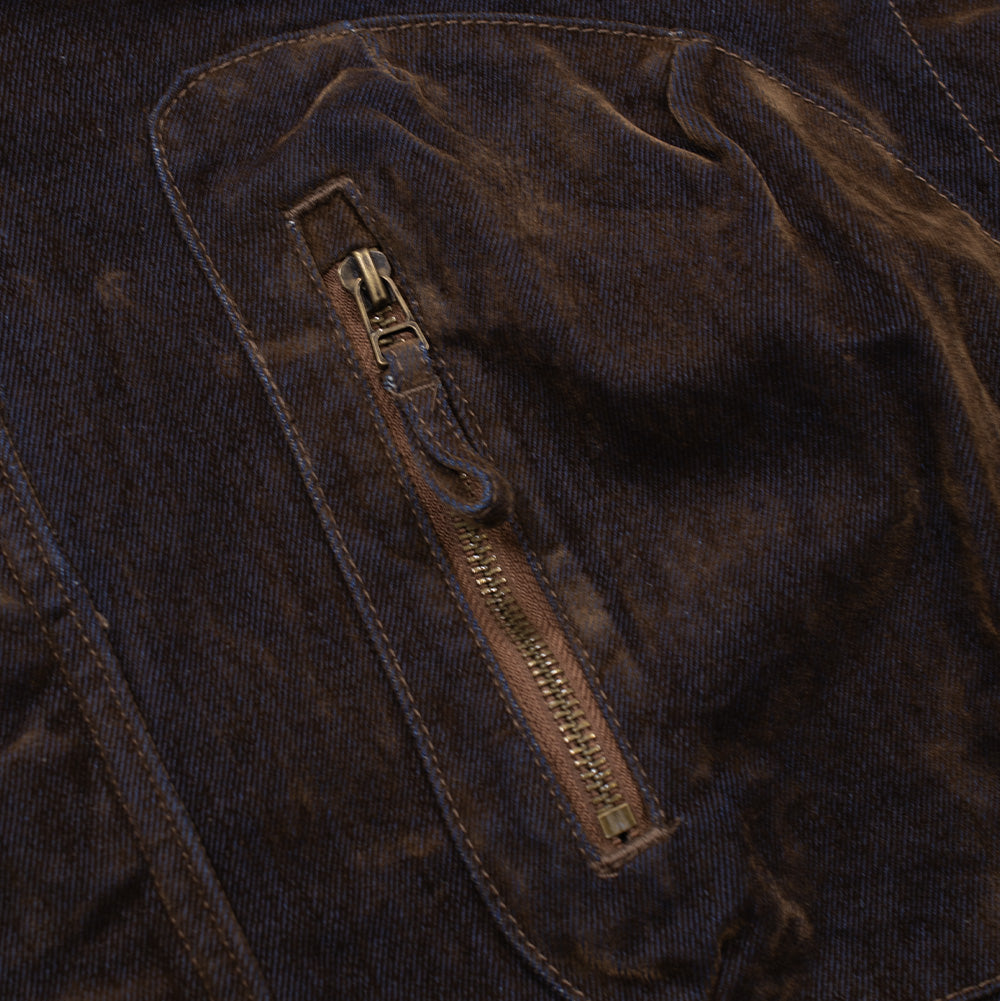 Macro perspective of the right chest pocket on the Brain Dead Flocked Denim Fireman Jacket, detailing the arched pocket silhouette, heavy brass zipper track with a matching denim pull tab, double-needle edge stitching, and the rich, mottled velvet texture of the brown flocking over blue denim.