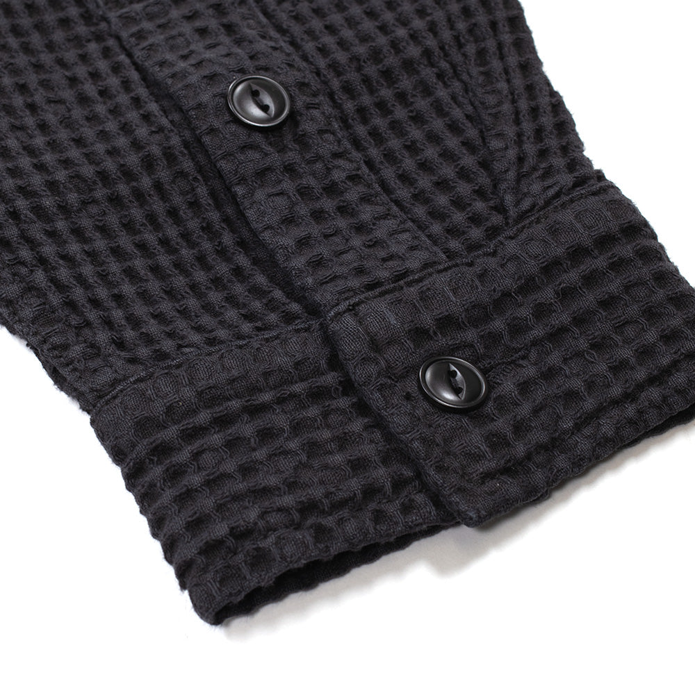 Macro studio perspective of the sleeve cuff on the Brain Dead Waffle Button Front Shirt in black, capturing the dual black buttons on the placket, reinforced cuff edge stitching, and the distinct, tactile honeycomb grid texture of the heavyweight dyed cotton waffle fabric.