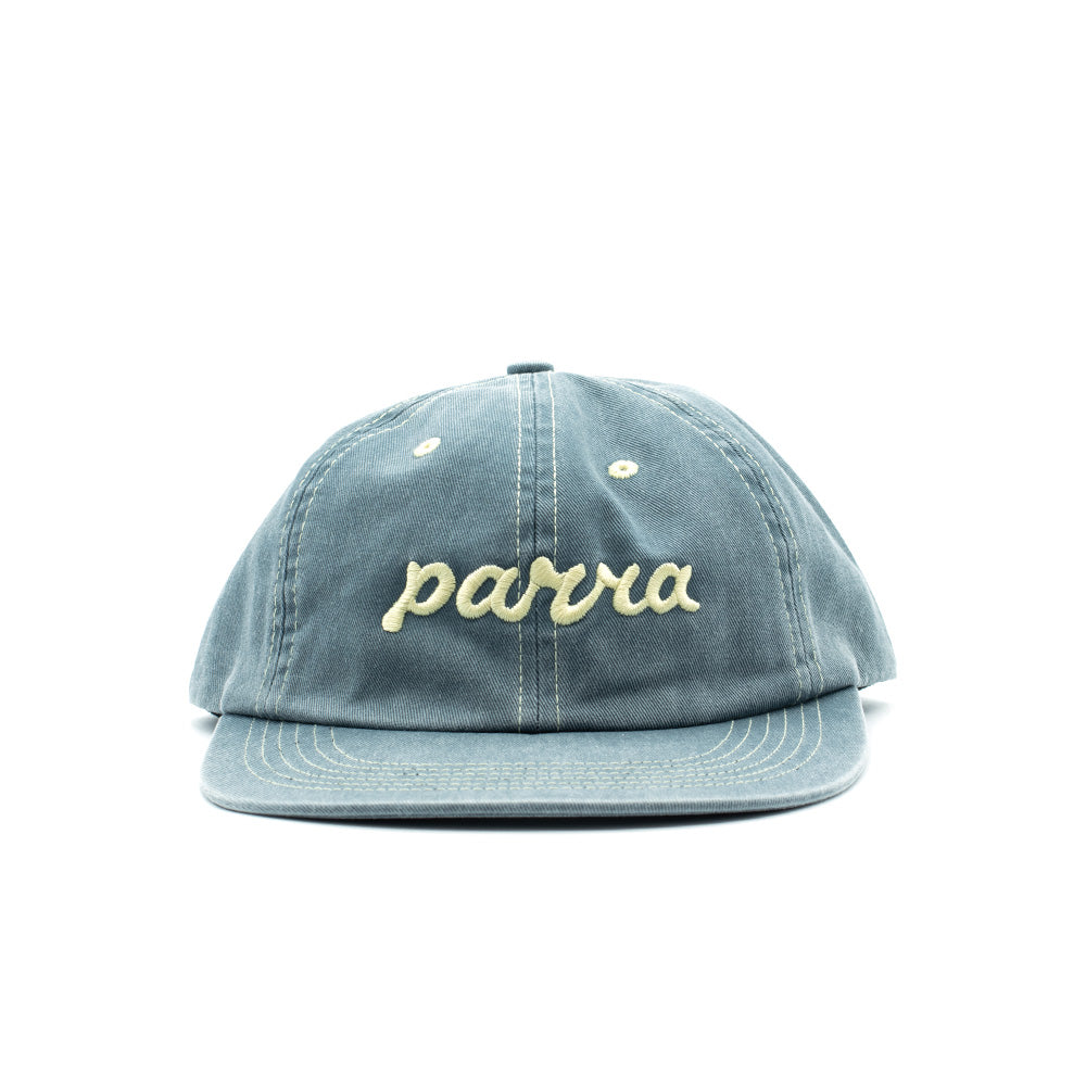 Front perspective studio view of the By Parra Lowercase Script Logo 6 Panel Hat in faded slate blue washed cotton twill, showcasing an unstructured low-profile polo cap silhouette with curved visor, contrast white eyelets and stitching, and cream-colored chain-stitch embroidered lowercase "parra" cursive script across the front crown.