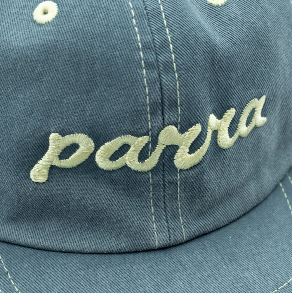 Detailed macro studio close-up of the front embroidery on the faded blue By Parra 6 Panel Hat, capturing the heavy, textured cream thread forming the cursive lowercase "parra" lettering against the soft, pre-washed diagonal cotton twill weave and stitched visor seam.