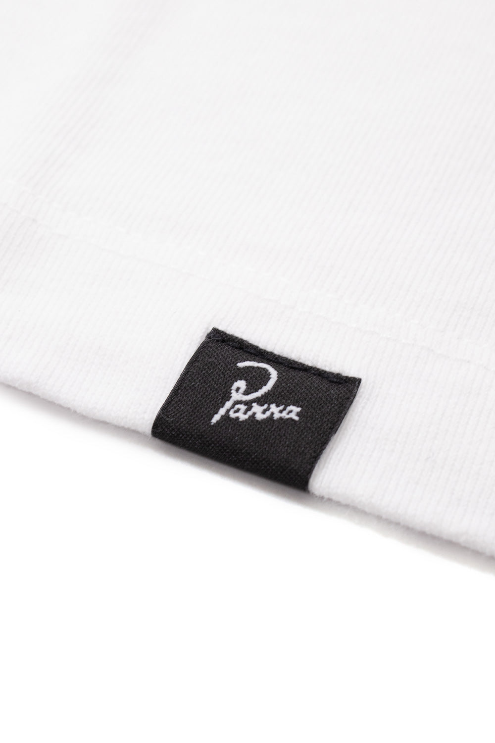 Extreme close-up detail shot of the bottom hem on the white By Parra T-Shirt, highlighting the clean double-needle stitch construction on the heavyweight 240g cotton jersey and the folded black woven side label featuring white embroidered cursive Parra script.