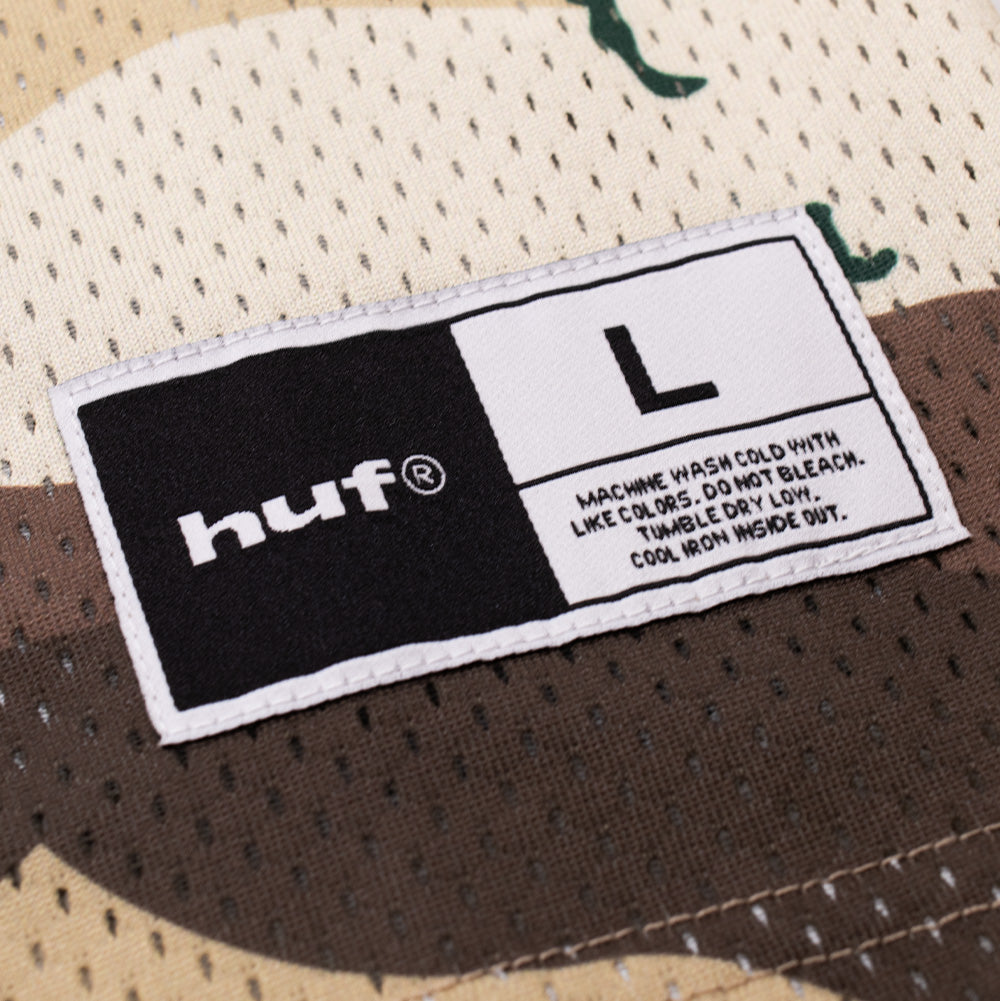 Macro studio perspective of the exterior woven athletic jock tag stitched onto the lower front hem of the Huf X Three 6 Mafia Football Jersey, showing the split black-and-white label with the white bold "huf®" logo, size "L", and care instruction lettering against the desert camo mesh background.
