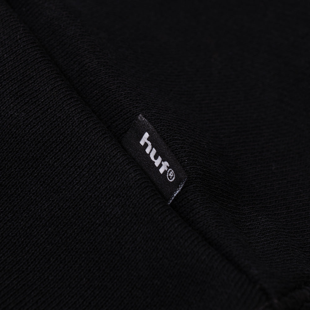 Detailed macro shot of the lower side seam on the Huf X Three 6 Mafia Heavyweight Hoodie, focusing on the small black woven HUF brand flag label with crisp white embroidered lettering, set against the thick heavyweight black brushed cotton fleece and ribbed hem stitching.