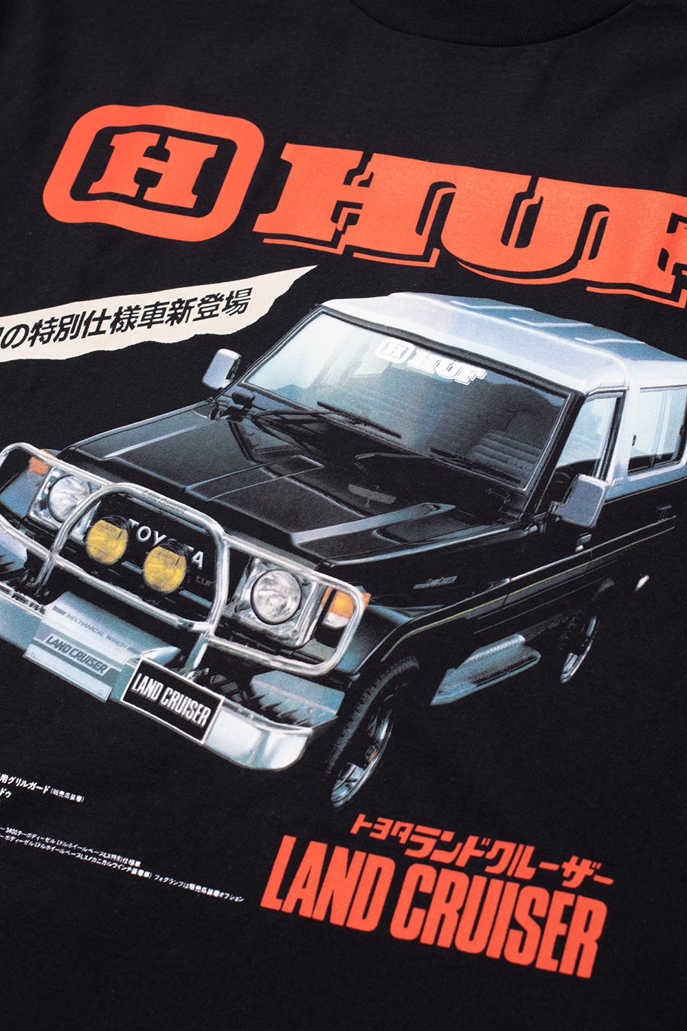 Detailed macro close-up of the front chest graphic on the Huf X Toyota Land Cruiser Turbo SS T-Shirt, highlighting the intricate vintage illustration of the 70-series 4x4 off-road vehicle with mechanical winch details, Japanese specification text banners, and orange-to-red gradient HUF and Land Cruiser branding on the black cotton jersey.