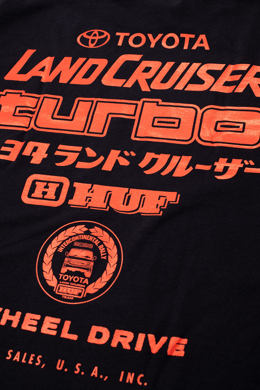 Studio macro perspective of the back graphic print on the Huf X Toyota Land Cruiser Turbo SS T-Shirt, detailing the textured, vibrant red-orange screen print of the classic "turbo" typography, Japanese Katakana translation, and circular laurel wreath rally emblem on the black textile.