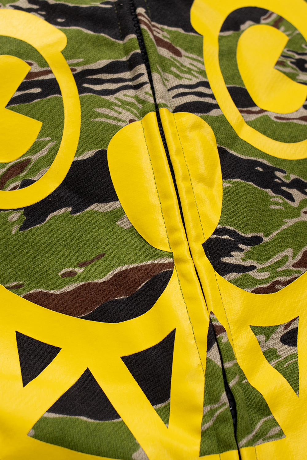 Close-up of IceCream Smiley full zip hood showing bright yellow printed face graphic over green tiger-stripe camouflage pattern.