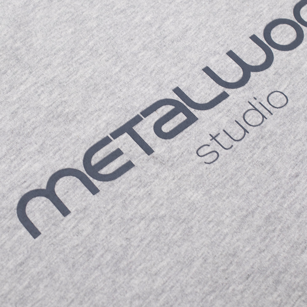 Detailed macro perspective of the center chest print on the Metalwood Studio Creature T-Shirt, displaying the smooth matte dark slate grey screen-printed "METALWOOD" modern rounded typeface stacked above "studio" on soft heather grey cotton jersey fabric.