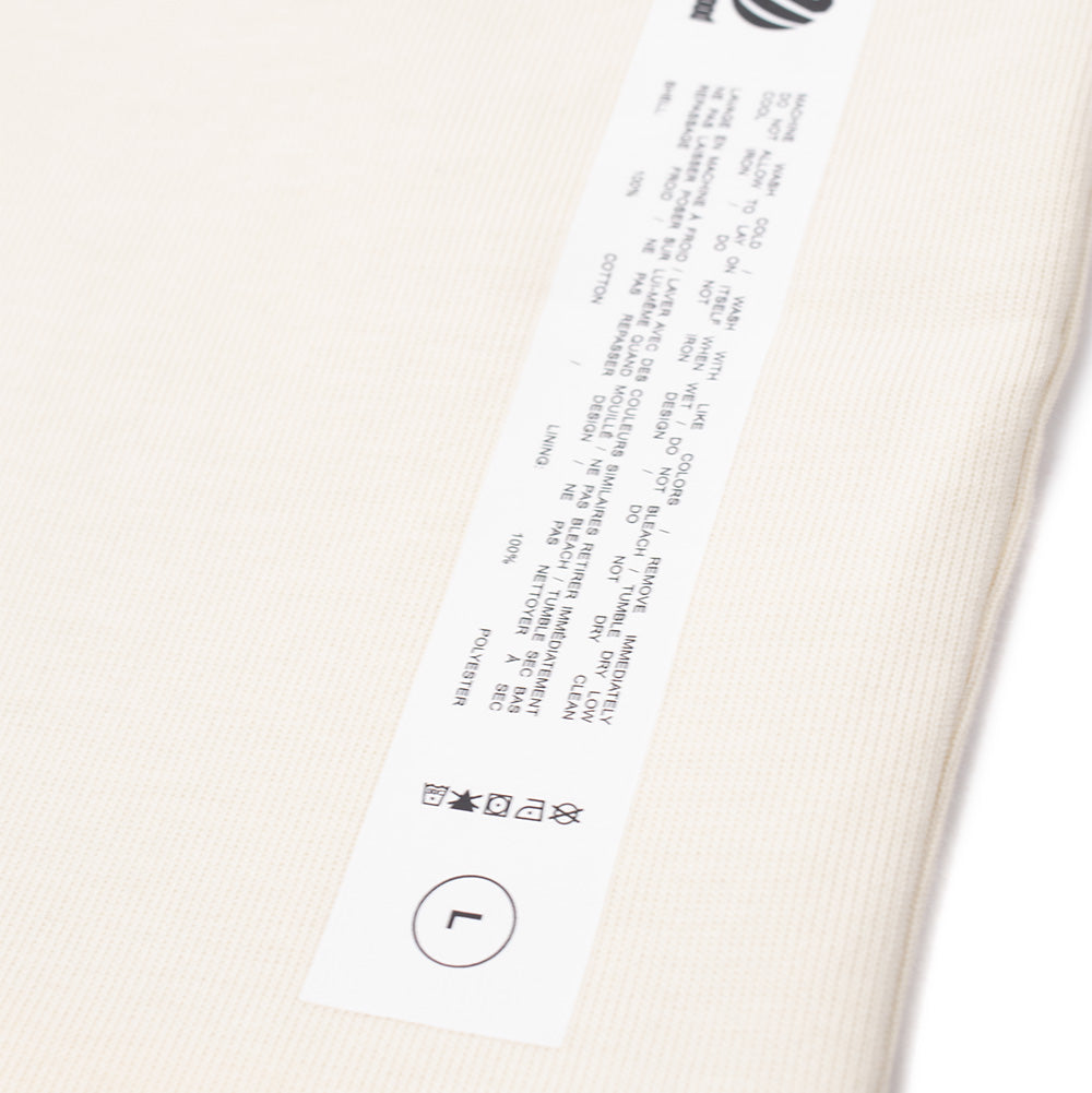 Macro studio view of the lower exterior label on the cream side of the Metalwood Studio Dual Face T-Shirt, showing the long rectangular white spec tag detailing bilingual washing instructions, material composition of 100% cotton shell and 100% polyester lining, circular "L" sizing, and the graphic Metalwood logo.