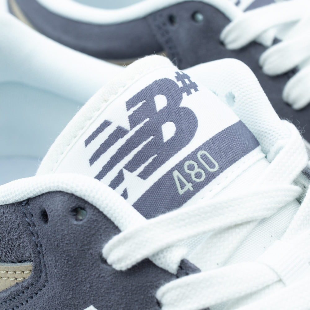 Macro close-up shot of the tongue label on the New Balance Numeric 480, focusing on the white woven label with blue-grey embroidered NB# logo graphic and clean "480" numerical model designation, framed by flat white athletic shoelaces and soft collar lining.