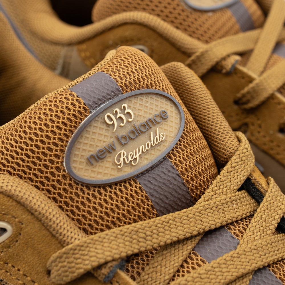 Macro close-up shot of the tongue on the New Balance Numeric Andrew Reynolds 933, highlighting the golden-brown waffle mesh fabric, flat woven laces, and the molded oval rubberized tongue label debossed with "933 new balance Reynolds" in white script lettering.