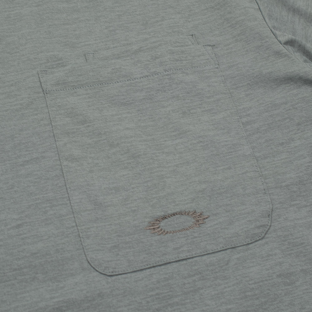 Macro studio view of the chest patch pocket on the Oakley FGL Scratch SS Tee, highlighting the double-stitched top hem with integrated pen slot and the tonal grey-green embroidery thread forming the jagged, scratch-style contour of the Oakley Ellipse emblem.