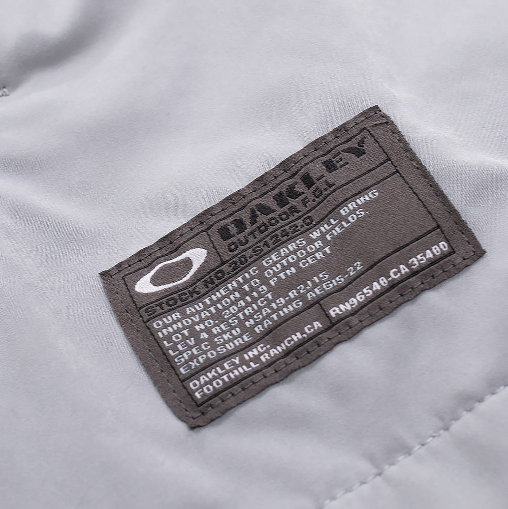 Macro studio view of the lower hem branding on the Oakley FGL Sector Jacket 6.7 in Winter Grey, focusing on the dark charcoal military-spec woven patch with white Oakley Ellipse icon, "OAKLEY OUTDOOR F.G.L." lettering, and authentic technical specification details stitched onto the matte shell fabric.