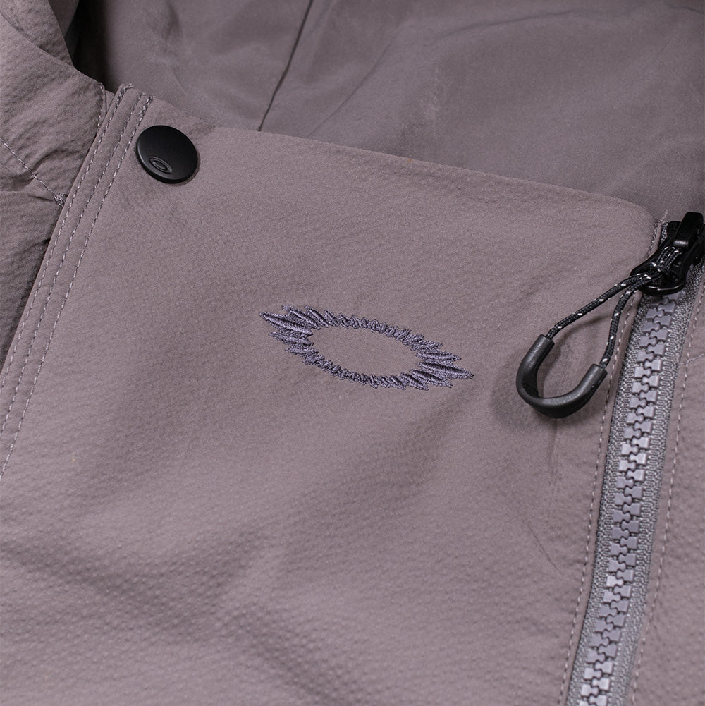 Detailed macro perspective of the high-collar closure on the Oakley FGL Stroke Anorak in Gray Wash, displaying the storm flap secured with a matte black snap button, asymmetrical chunky plastic zipper track with a cord loop puller, and tonal charcoal embroidery outlining the radiating scratch-style Oakley Ellipse emblem.