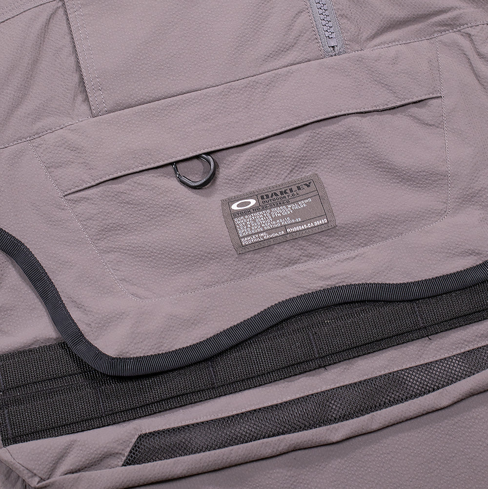 Macro studio view of the central chest utility pouch on the Oakley FGL Stroke Anorak, showing the curved protective flap with D-ring attachment, horizontal zipper pocket, dual black modular webbing gear loops, bottom mesh aeration insert, and a dark rectangular military-spec tag with white Oakley branding and technical product codes.