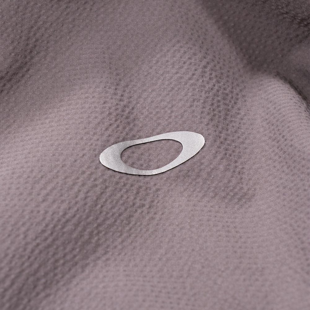 Extreme macro close-up of the rear shoulder branding on the Oakley FGL Stroke Anorak, displaying the reflective silver heat-transfer Oakley Ellipse icon seamlessly bonded onto the fine, weather-resistant micro-texture of the Gray Wash woven technical fabric.