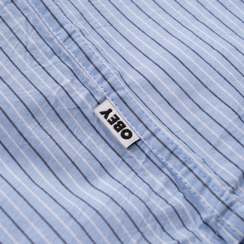 Studio macro perspective of the lower side seam on the Obey Billy Woven Shirt, showing the neat double-needle stitching on the curved hem and the small white woven OBEY brand flag label with black block lettering.