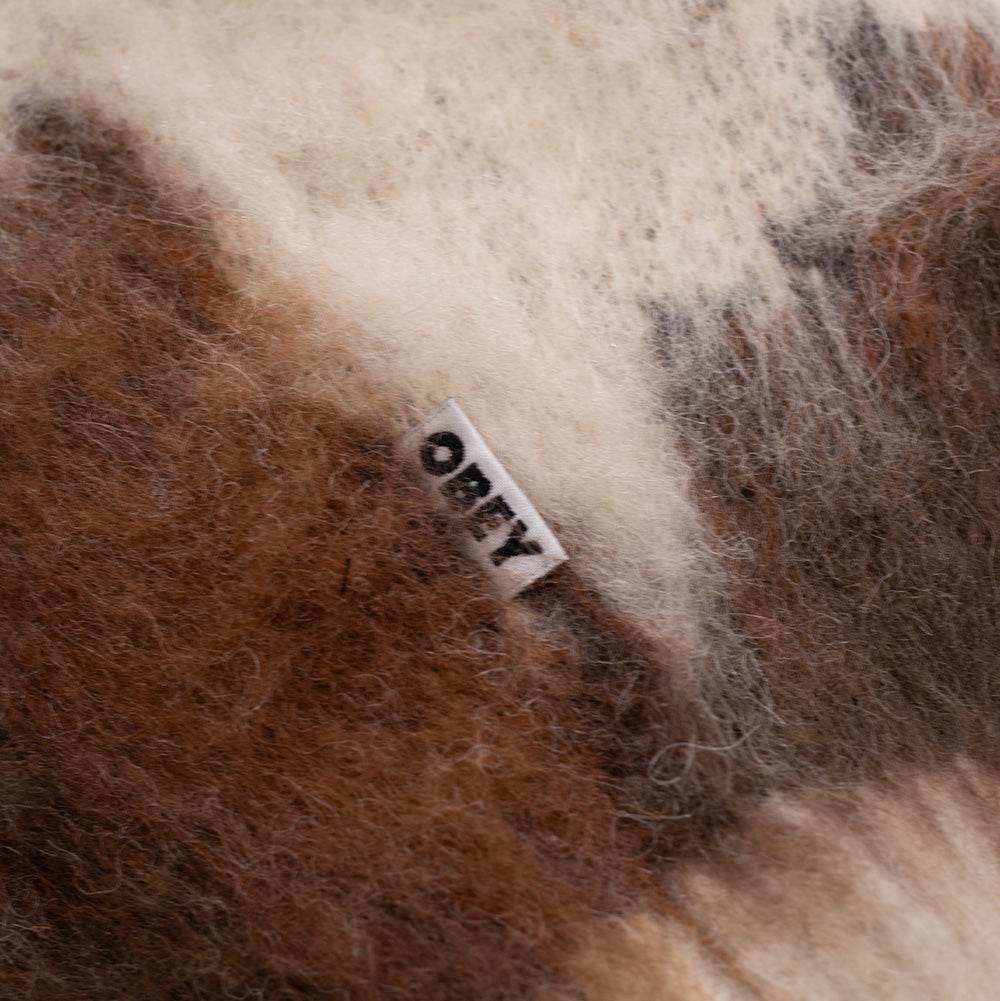 Extreme macro perspective of the side seam on the Obey Cow Pattern Zip Sweater, showcasing the fluffy, hairy fibers of the brushed yarn and the small white woven OBEY logo tag with black lettering.