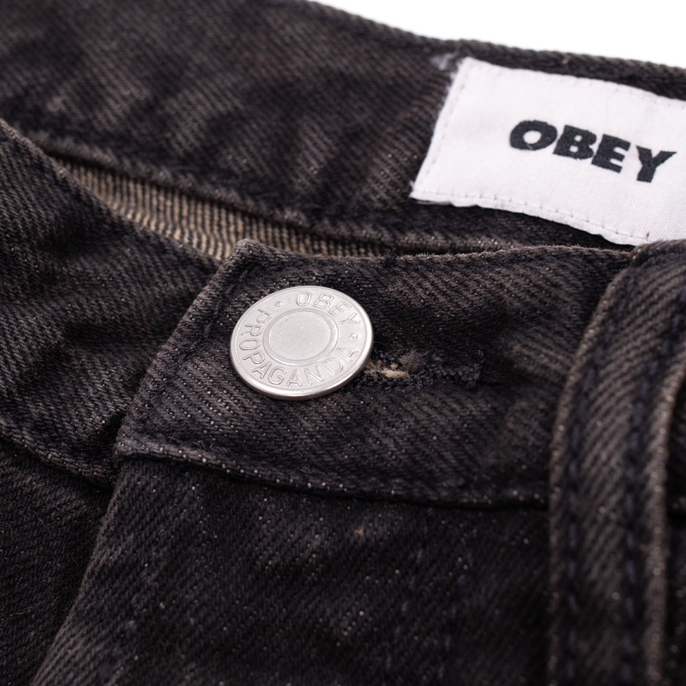 Macro studio close-up of the waistband closure on the Obey Studded Boxcar Denim, detailing the silver metallic donut shank button stamped with "OBEY PROPAGANDA", heavy twill denim diagonal weave with rust undertones, and the interior white woven OBEY brand tag.