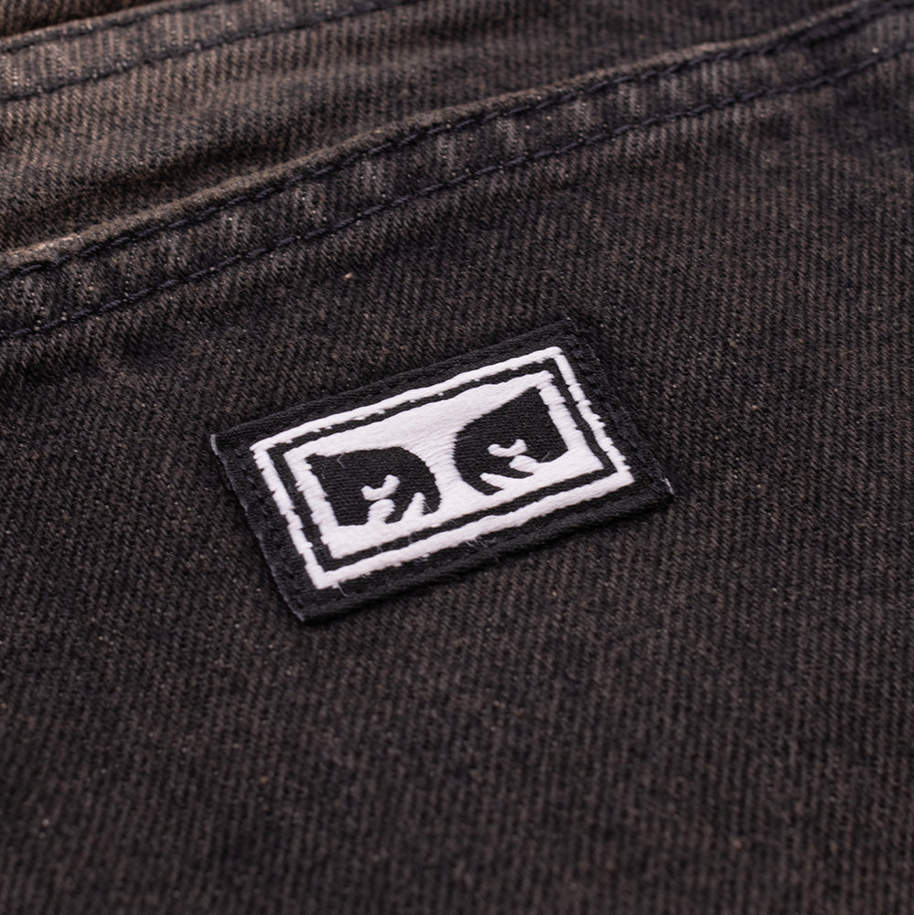 Studio macro perspective of the back pocket on the Obey Studded Boxcar Denim, highlighting the miniature black and white woven rectangular label with the iconic Obey Eyes motif stitched cleanly onto the washed faded black denim twill.