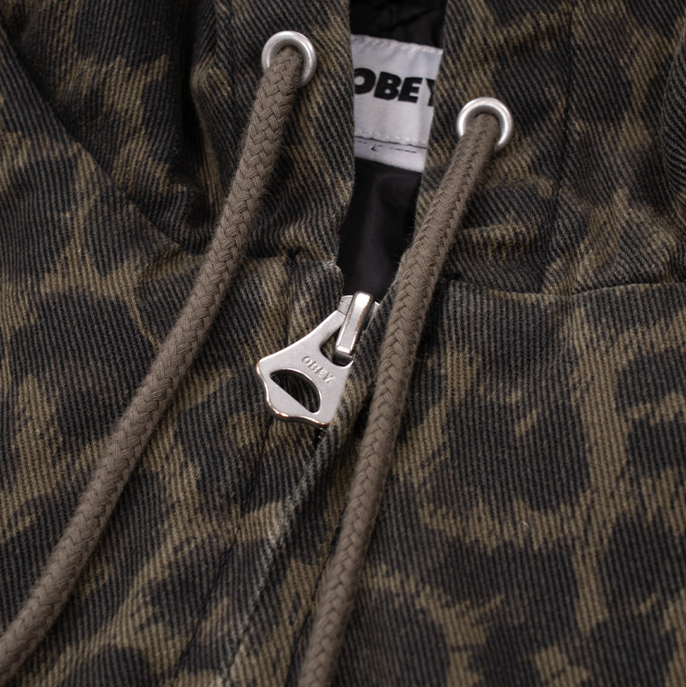 Macro studio close-up of the front zipper and collar of the Obey Studded Zip Up Jacket in Dark Leopard, highlighting the thick silver-tone zipper featuring an engraved OBEY logo pull, matching braided olive drawstrings threaded through silver eyelets, and the leopard pattern printed on heavy cotton twill.