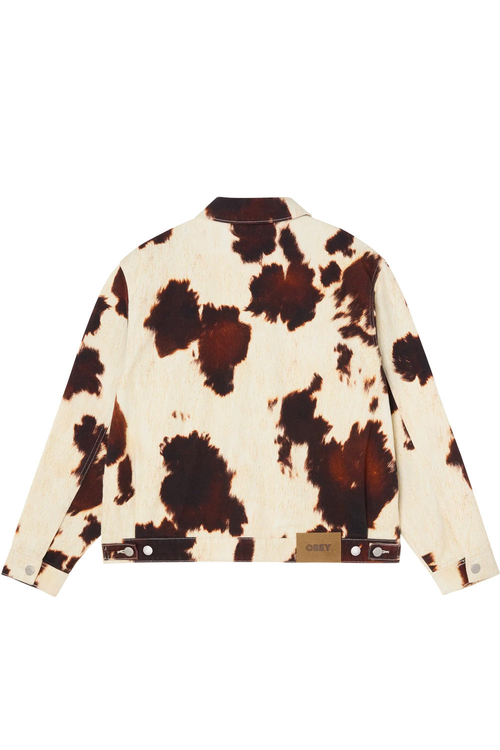 Back view of the relaxed fit Obey Workshop denim jacket in brown and cream cow print, displaying straight-cut shoulder paneling, adjustable button waist tabs, and an embossed brown suede leather Obey logo patch on the rear waistband.