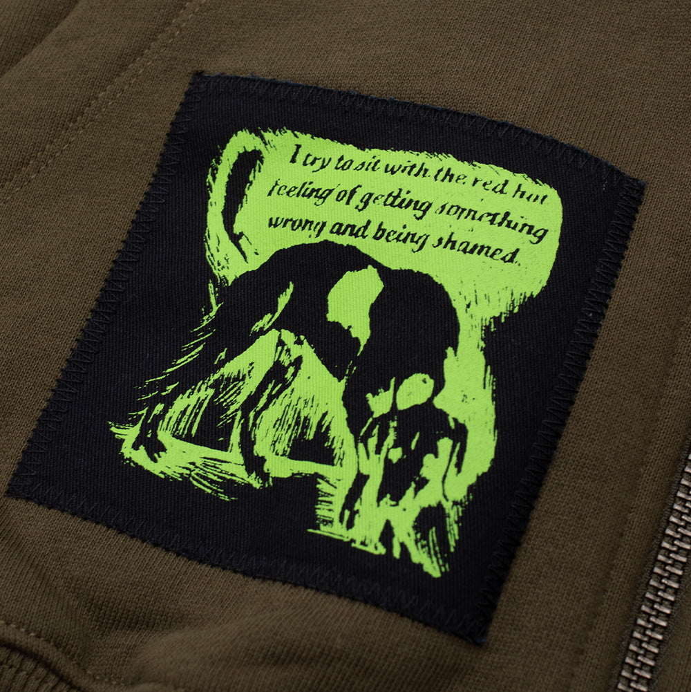 Detailed macro shot of the lower front pocket patch on the Token Carly Zip Hoodie, showcasing a neon green and black block print of a dog alongside text that reads "I try to sit with the red hot feeling of getting something wrong and being shamed."
