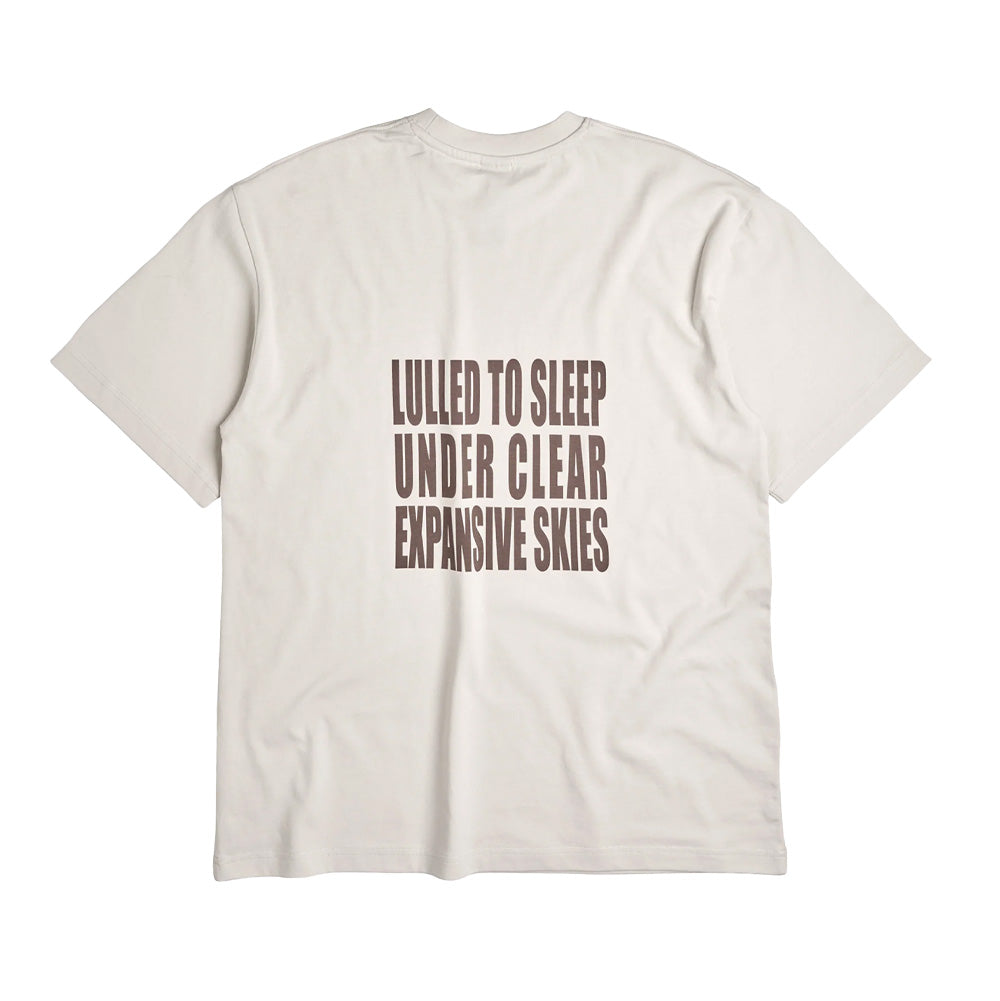 Back flat lay view of the off-white Token Expansive Skies T-Shirt, showing a clean streetwear silhouette centered with a large four-line typography print in dark brown condensed sans-serif block capitals reading "LULLED TO SLEEP UNDER CLEAR EXPANSIVE SKIES".