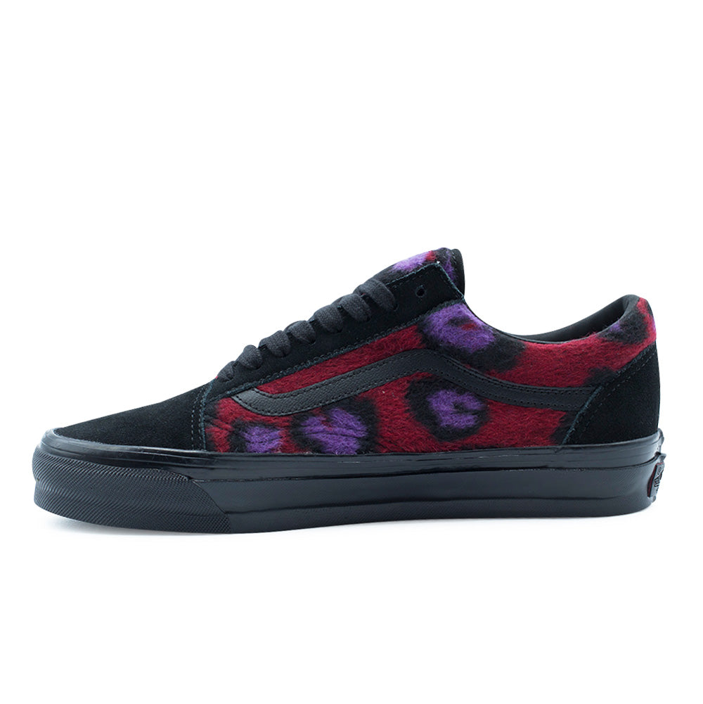 Medial side studio profile of the Vans LX Old Skool shoe, showing the continuous fuzzy fleece leopard pattern in crimson red, violet purple, and jet black, stitched full-grain black leather sidestripe, Osnaburg-reinforced black foxing tape, and black suede heel wrap.