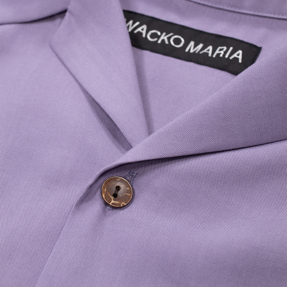 Macro studio close-up of the open collar and top button closure on the purple Wacko Maria 50's Shirt, highlighting the sharp notch lapel construction, finely textured drape of the lavender lyocell fabric, natural marbled brown coconut-style two-hole button, and the black woven "WACKO MARIA" neck label.