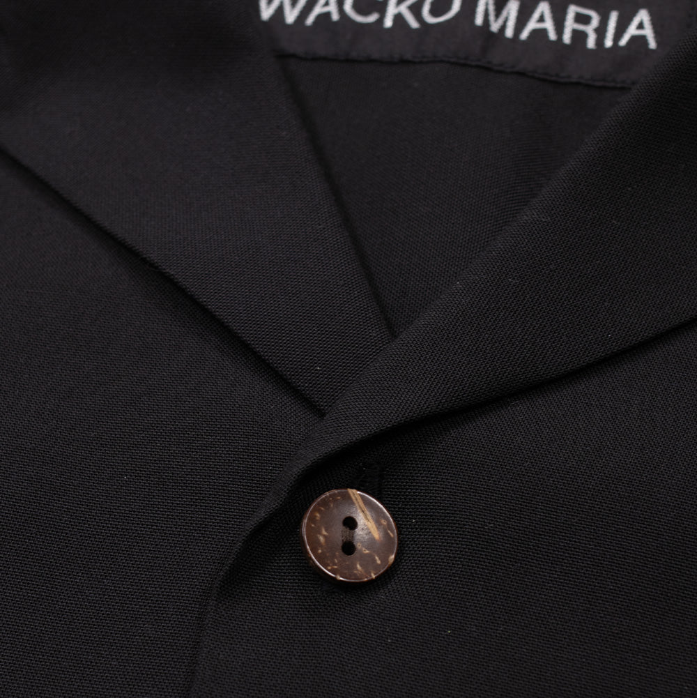 Macro studio close-up of the camp collar and upper button on the Wacko Maria 50's Shirt, highlighting the notch lapel construction, finely textured matte woven charcoal twill fabric, natural brown carved wooden button with contrast marbling, and the white woven "WACKO MARIA" brand neck tag.