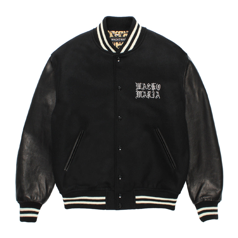 Front flat lay studio view of the black Wacko Maria Leather Varsity Jacket Type 3, featuring a snap-button front closure, double-welt leather hand pockets, striped knit ribbing at the stand collar, hem, and cuffs, with white embroidered gothic "WACKO MARIA" lettering on the left chest and a glimpse of the leopard-print lining.