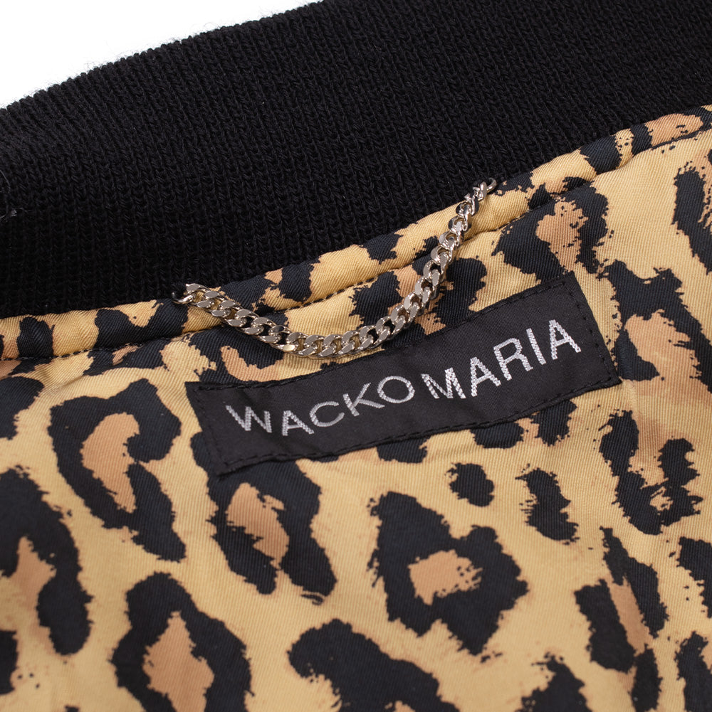 Macro studio view of the inner neckline of the Wacko Maria Leather Varsity Jacket, displaying the woven black rectangular "WACKO MARIA" brand label stitched onto the leopard satin lining below a polished silver curb-chain hanging loop.