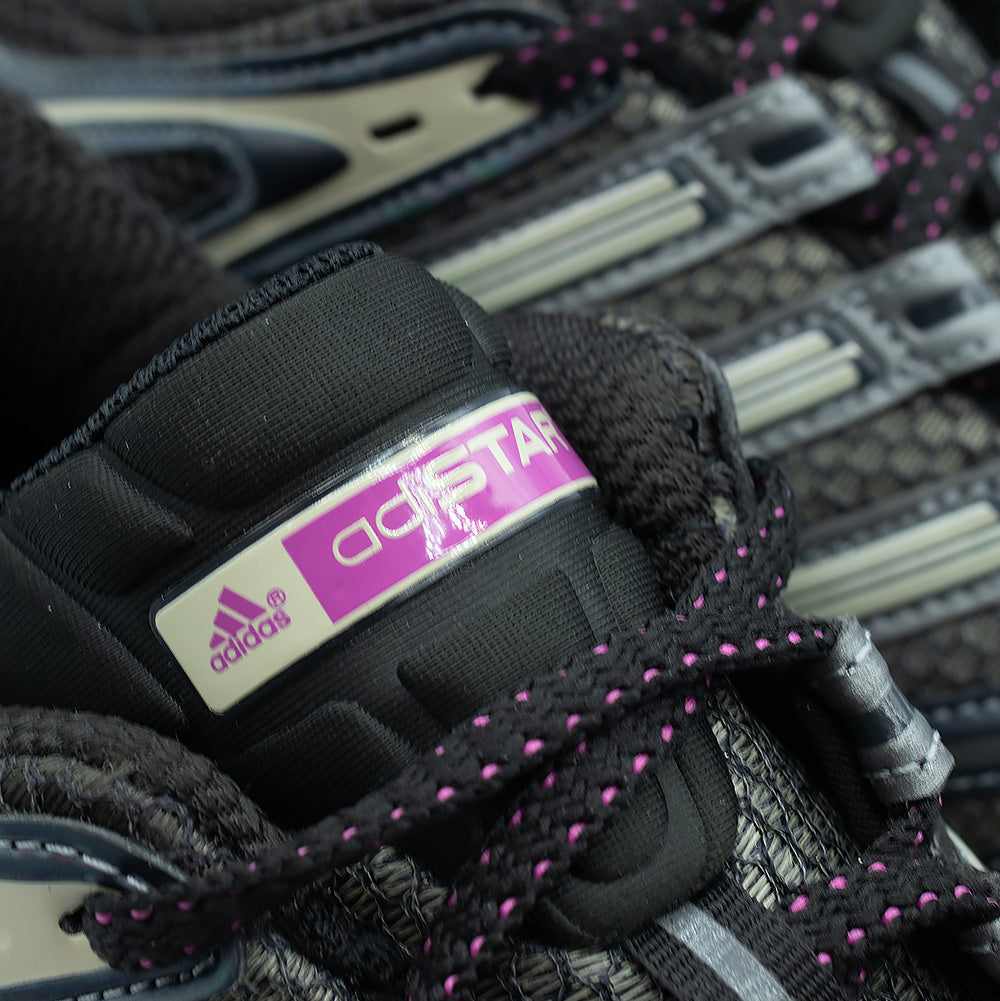 Macro close-up shot of the tongue branding on the adidas Adistar Control 5, highlighting the padded black fabric tongue, black and fuchsia speckled laces, and the molded tongue badge with clean white and fuchsia adidas performance logo and "adiSTAR" typography.