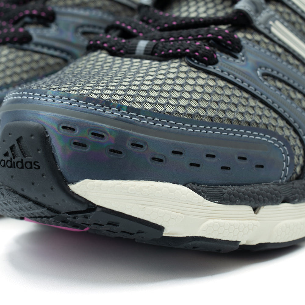Detailed macro shot of the toe cap on the adidas Adistar Control 5, displaying the pearlescent iridescent synthetic toe guard with ventilation punch-outs, double-stitched seams, breathable honeycomb mesh underlay, and the textured black rubber toe bumper embossed with the adidas logo.