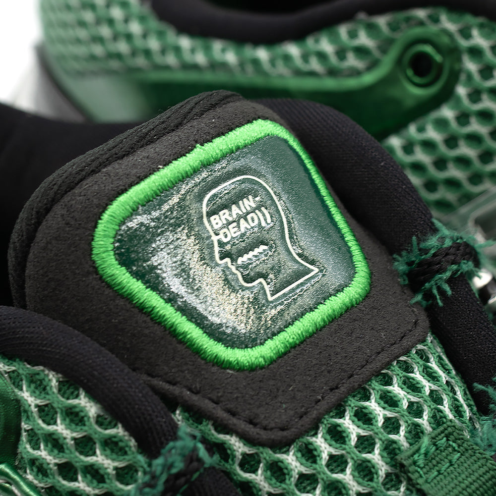 Macro close-up shot of the tongue badge on the adidas x Brain Dead Barricade V, highlighting the embossed white Brain Dead profile head logo inside a glossy dark green badge bordered by vivid neon green satin embroidery, set over breathable open-cell mesh.