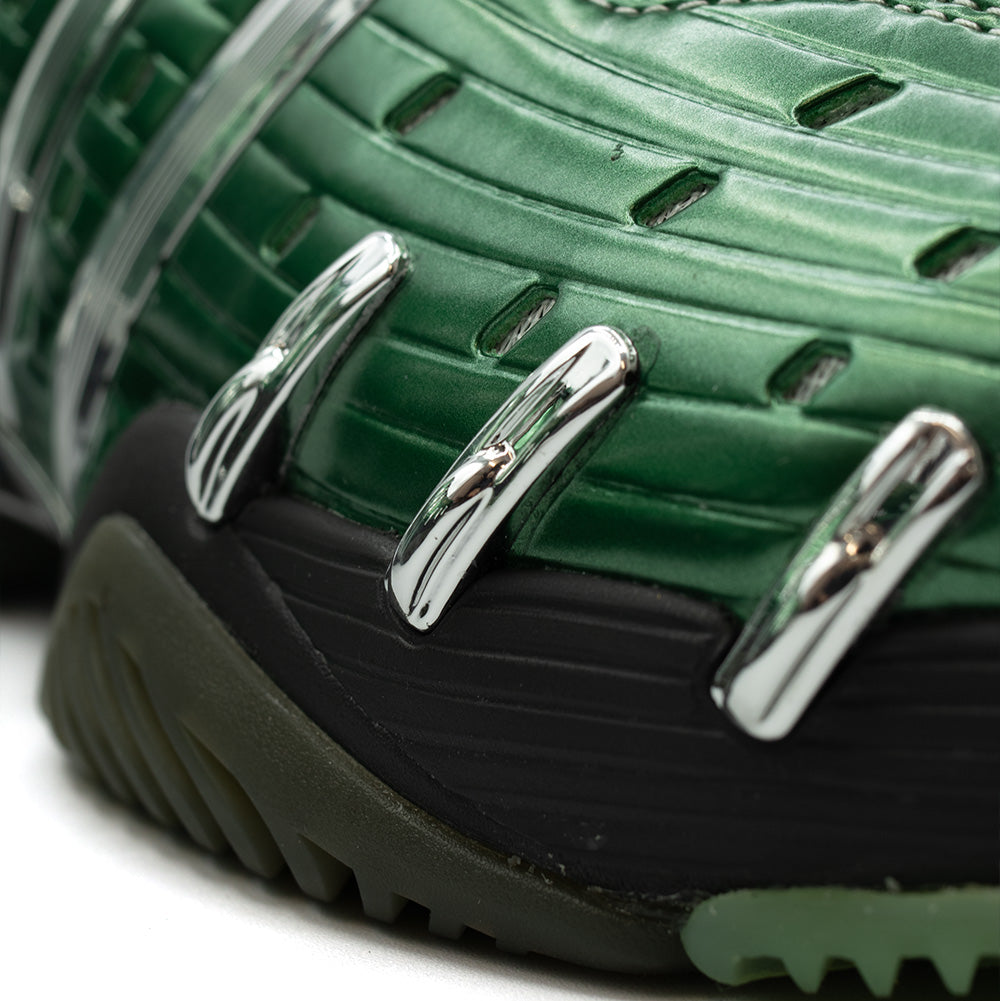 Extreme macro close-up of the lateral forefoot on the adidas x Brain Dead Barricade V, displaying the metallic emerald green ribbed shell, ventilation cutouts, shiny chrome claw accents, and textured black lateral outsole wrap.