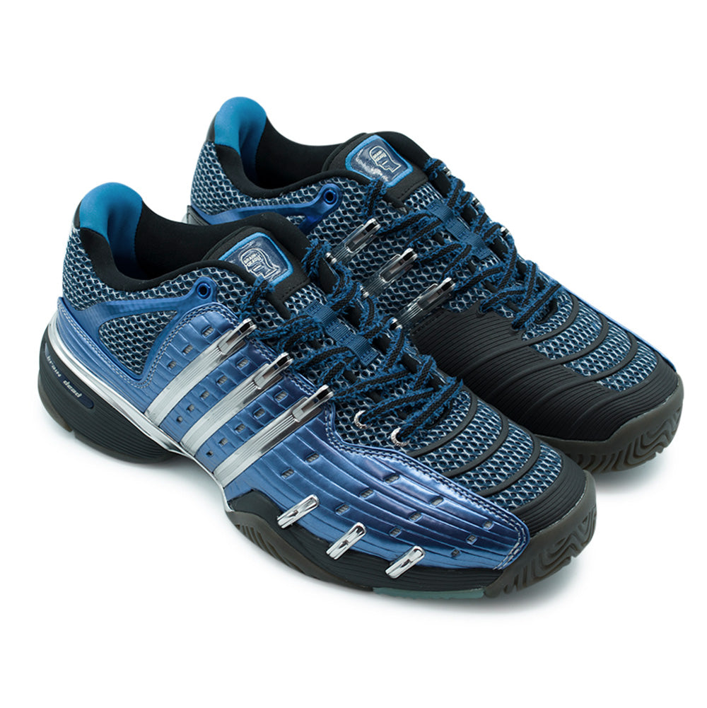 Three-quarter studio perspective of the adidas x Brain Dead Barricade V sneakers, highlighting the heavy-duty 2008 court chassis, shiny chrome midfoot eyelet anchors, ribbed protective forefoot panels, two-tone laces, and molded blue tongue badges.