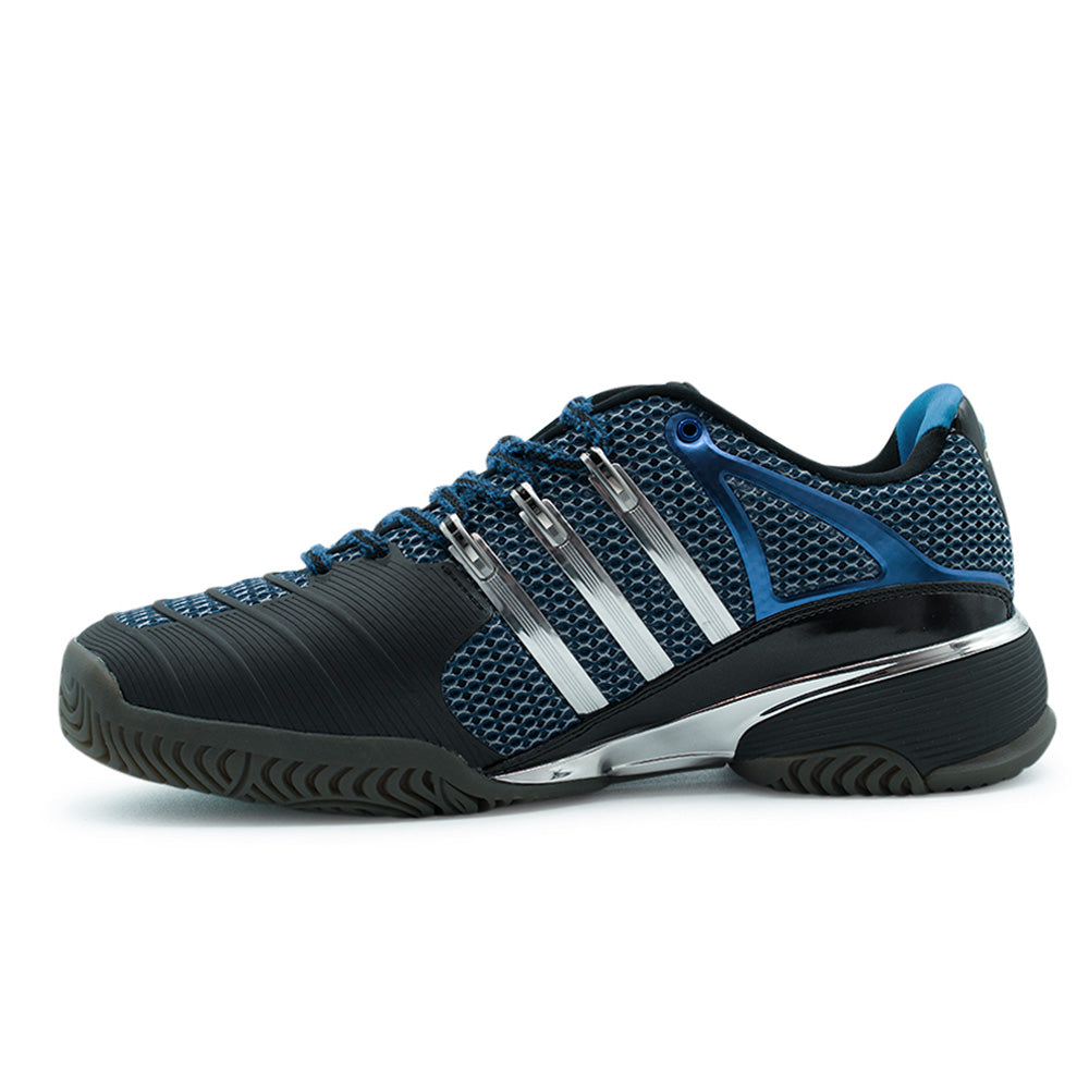 Medial side studio profile of the adidas x Brain Dead Barricade V, showing the extensive matte carbon-black textured rubber toe-drag protection guard, breathable blue sandwich mesh midfoot underlay, polished silver three stripes, and chrome midfoot stabilization plate.